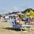 Woman at the beach relaxes by reading a gossip magazine. On the background a peddler is walking. Royalty Free Stock Photo